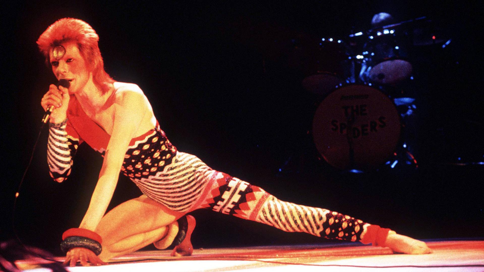 Der Sänger Davis Bowie hockt ei einem Konzert in einem bunten Ganzkörperanzug auf der Bühne.