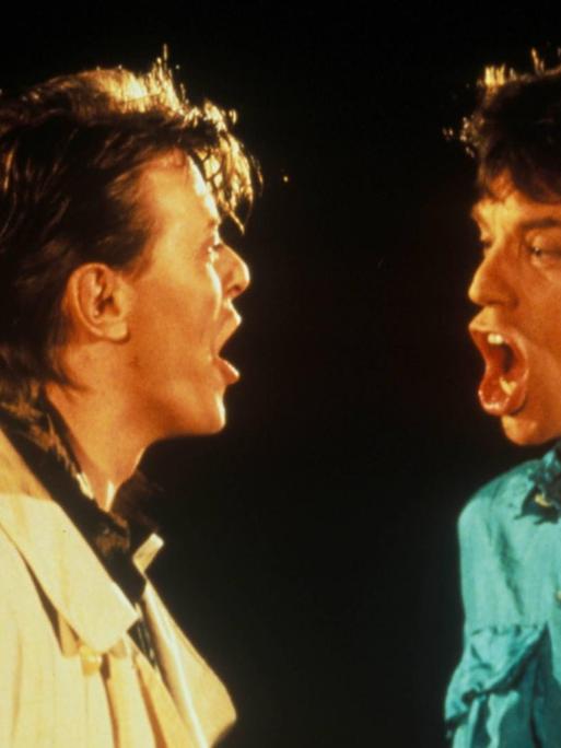 David Bowie links in heller Jacke und Mick Jagger rechts in türkisem Hemd sind im Profil zu sehen. Beide haben den Mund offen und schauen sich an, während sie "Dancing in the street" singen.