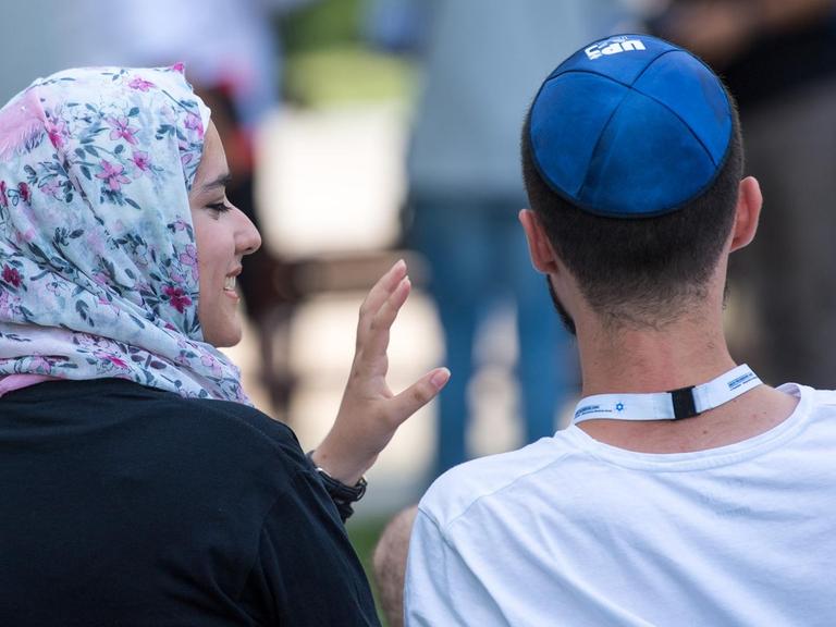 Eine junge Muslima mit Kopftuch und ein junger jüdischer Mann mit Kippa unterhalten sich miteinander.