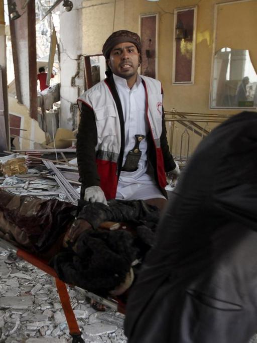 Drei Männer tragen das Opfer auf einer Bahre aus der zerstörten Halle, sie werden von einem Arzt begleitet. Andere Menschen schauen von der Seite zu. Der Boden ist mit Trümmern übersät.