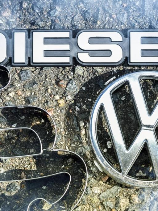 Ein VW-Logo, Paragrafen- und Dieselzeichen liegen auf der Straße.