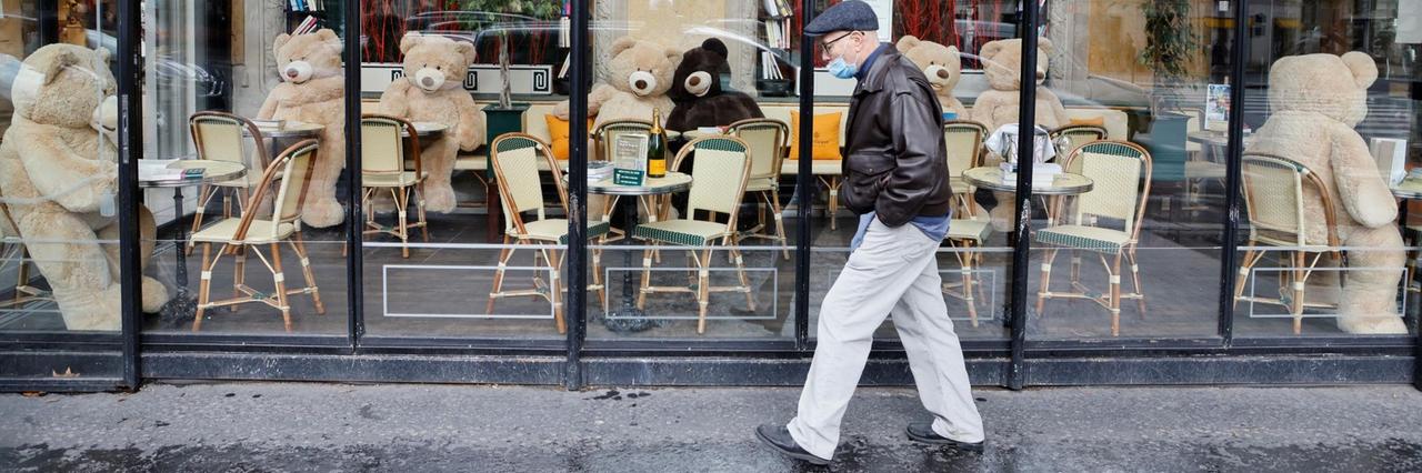 Ein Mann mit Atemschutzmaske geht an einem Café vorbei. Dieses ist geschlossen. An den Tischen im Café sitzen überdimensionale Teddybären. Ein Mann mit Atemschutzmaske geht an einem Café vorbei. Dieses ist geschlossen. An den Tischen im Café sitzen überdimensionale Teddybären.