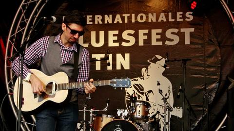 Ein Mann mit Kinnbart und Sonnenbrille steht vor dem Schriftzug "Bluesfest Eutin" auf einer Bühne und spielt akustische Gitarre.