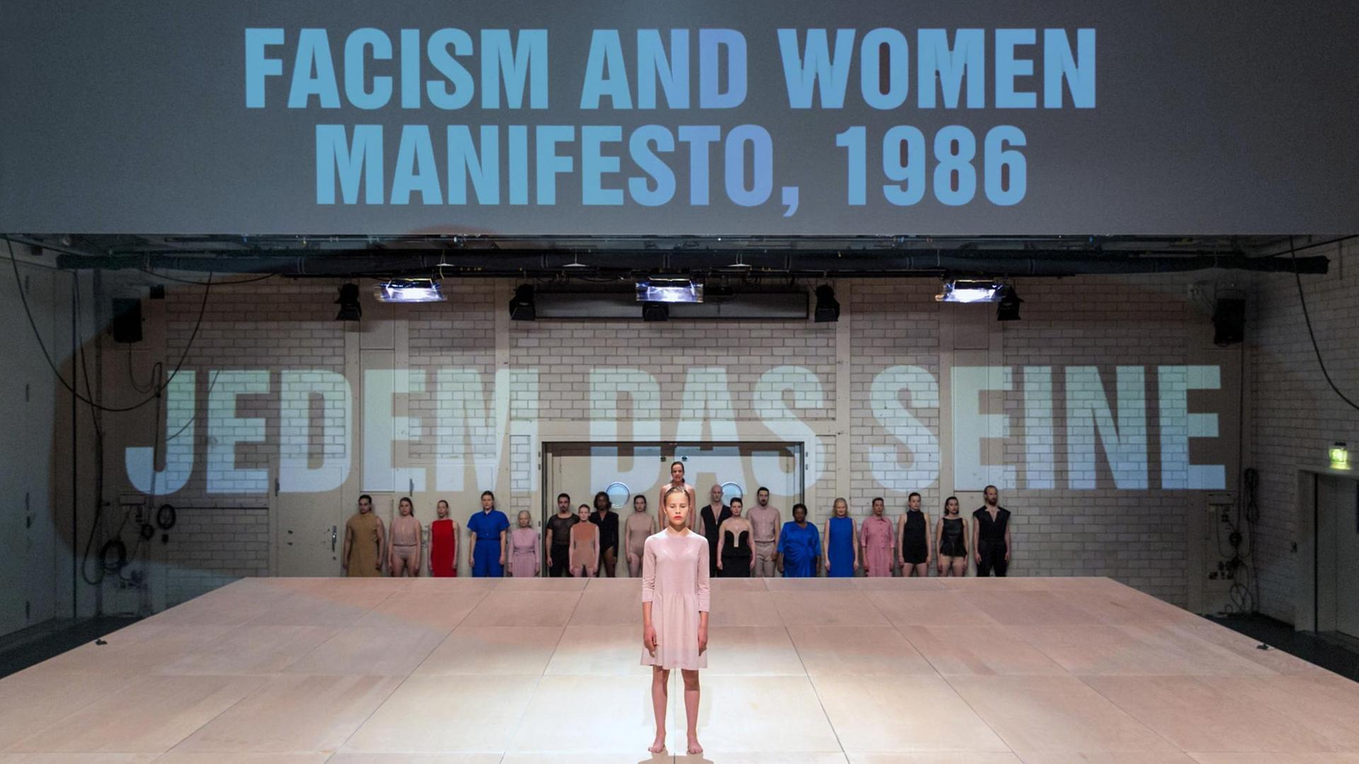 Eine Frau steht auf einer Theaterbühne, im Hintergrund stehen die Wörter: "Facism and Woman Manifesto, 1986" und "Jedem das seine"