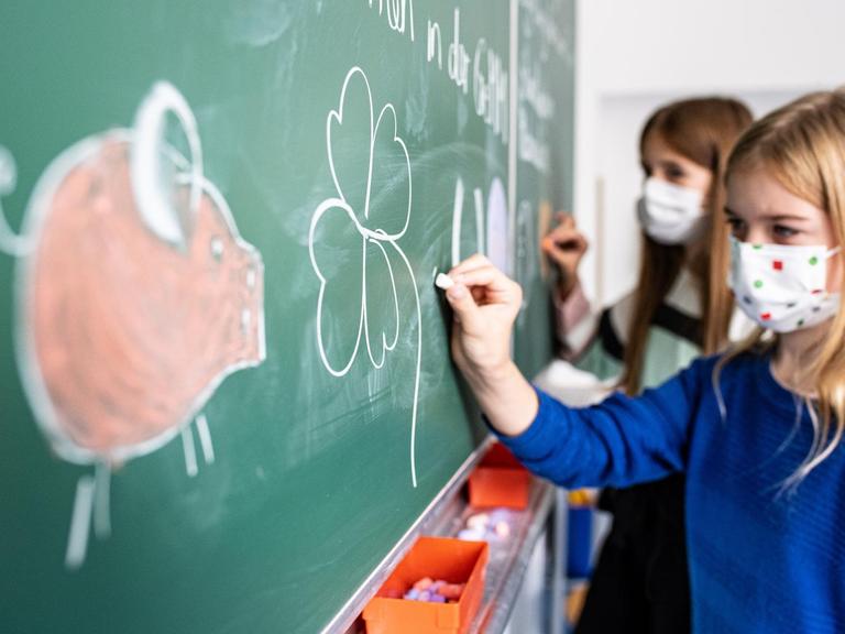 Zwei Schülerinnen mit Schutzmaske malen ein Schwein, Kleeblatt, Hufeisen und einen Luftballon an die Tafel in einem Klassenzimmer.