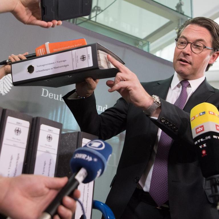 Scheuer hält einen Aktenordner hoch und zeigt mit dem Zeigerfinger darauf. Um ihn herum werden Mikrofone der Sender gehalten.