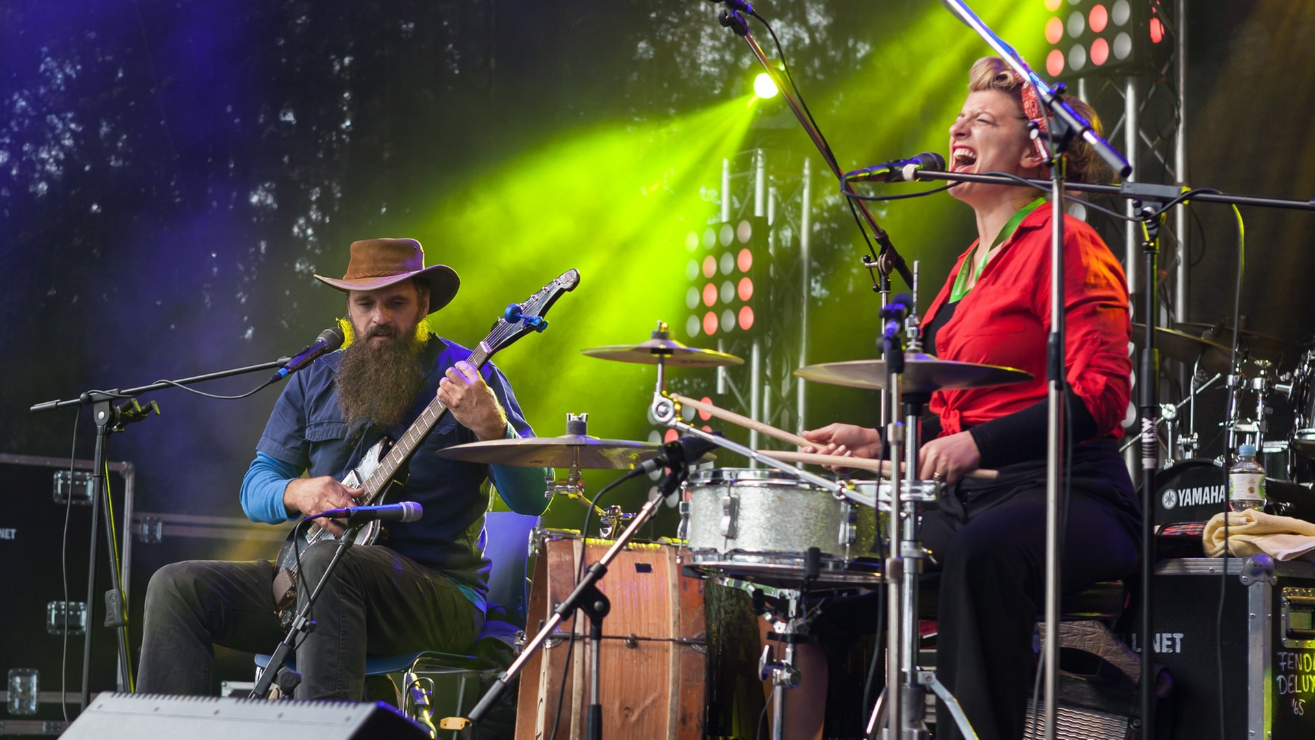 Eine Frau in roter Bluse spielt Schlagzeug und singt. Neben ihr sitzt ein Mann und spielt Gitarre.