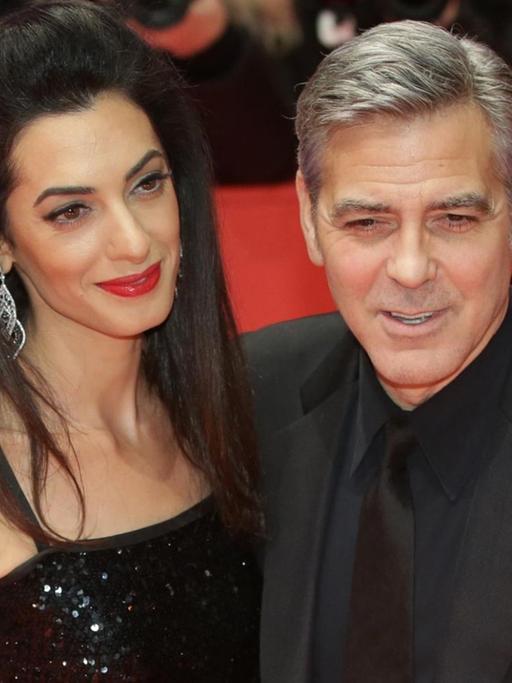 Clooney und Amal lächeln auf dem roten Teppich engumschlungen in die Kameras. Er trägt einen schwarzen Anzug, sie ein schwarzes schulterfreies Kleid.
