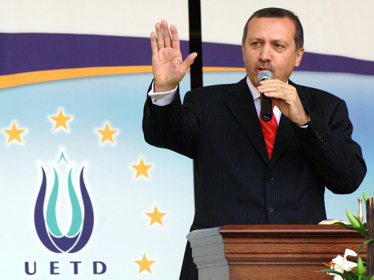 Erdogan spricht an einem Rednerpult in ein Mikrofon, das er mit der linken Hand hält; mit rechts gestikuliert er. Im Hintergrund das Logo der Union Europäisch-Türkischer Demokraten UETD.