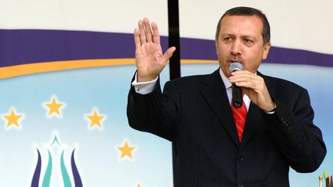 Erdogan spricht an einem Rednerpult in ein Mikrofon, das er mit der linken Hand hält; mit rechts gestikuliert er. Im Hintergrund das Logo der Union Europäisch-Türkischer Demokraten UETD.