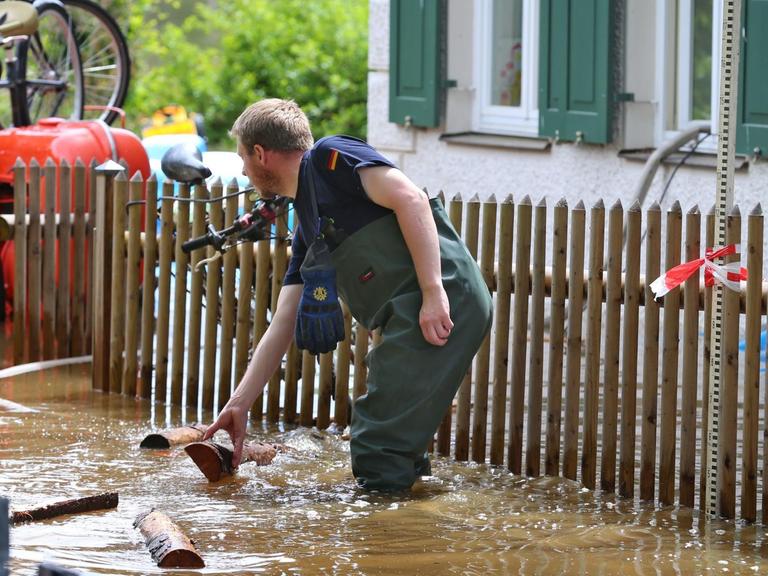 Ein Mitarbeiter des Technischen Hilfswerks steht knietief im schlammigen Wasser, hinter ihm ein Gartenzaum und ein Wohnhaus. Gegenstände treiben durch das Wasser.