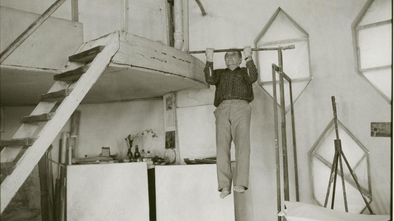 Die Fotografie "Sohn des Architekten Melnikow" von Helga Paris, aus der Serie "Moskau" (1991/92). Ein älterer Mann macht Klimmzüge an einer provisorischen Querverstrebung an einer hölzernen Treppe.