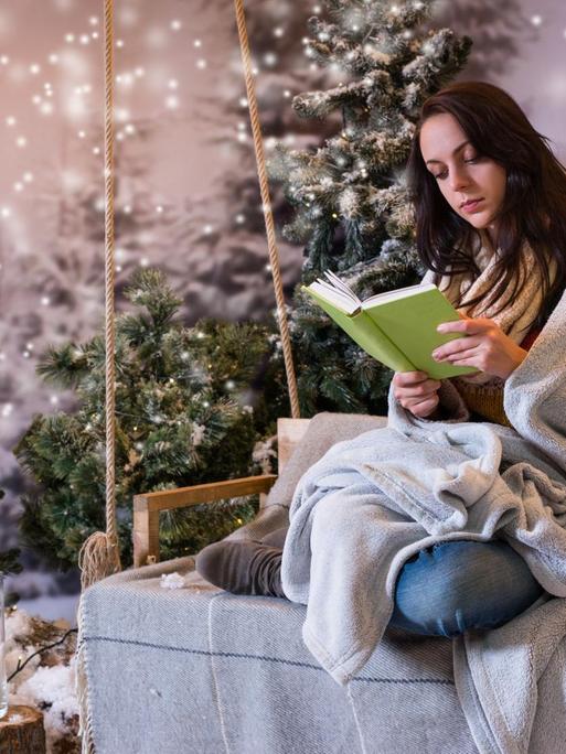 Eine junge Frau sitzt vor romantischer Weihnachtskulisse und liest ein Buch.