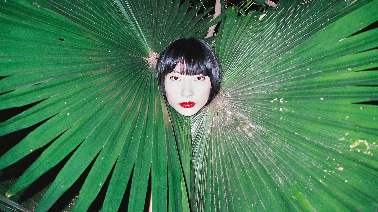 Eine Frau schaut in Mitten zweier Palmenblätter den Fotografen an, "Untitled, 2015" von Ren Hang.