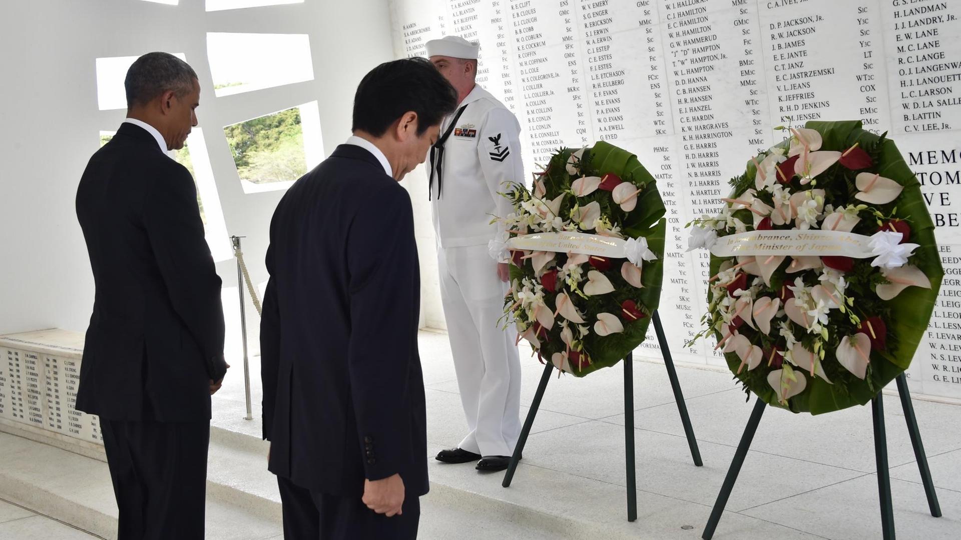 US-Präsident Obama und Japans Ministerpräsident Abe haben in der offizoiellen Gedenkstätte in Pearl Harbor auf je einem Ständer Kränze abgelegt. Im Hintergrund sind auf einer weißen Marmorwand die Namen der Gefallenen eingraviert.