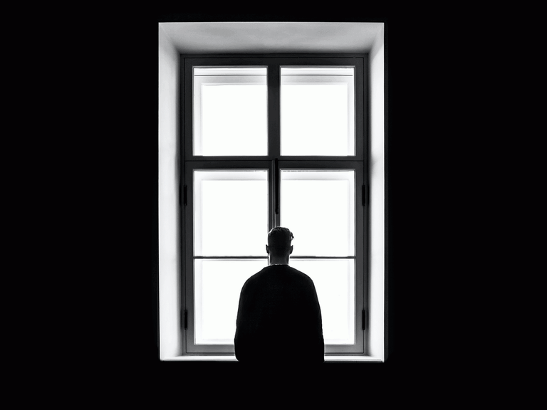 Ein Mann steht mit dem Rücken zur Kamera und schaut aus einem Fenster. Die Wände sind schwarz.