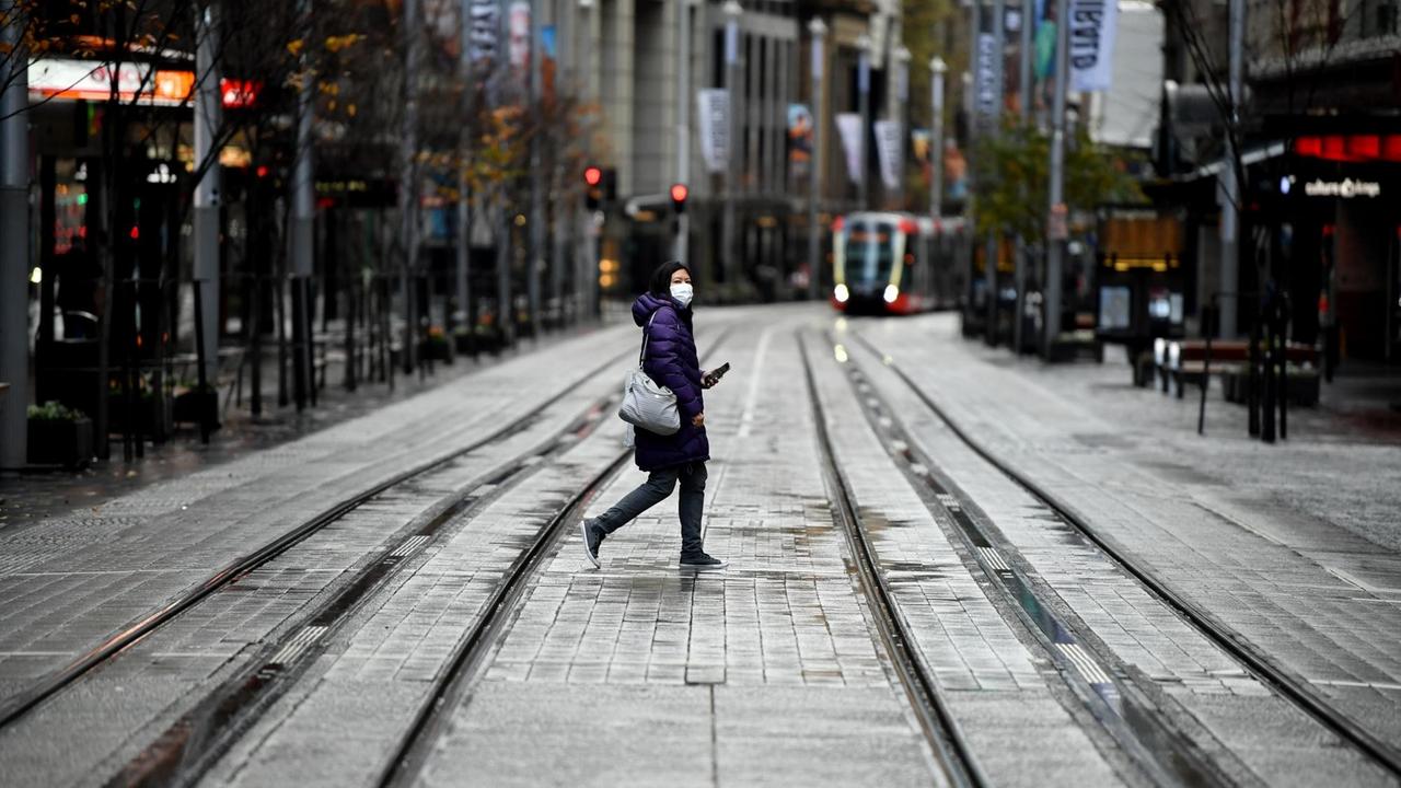 Eine Frau mit Maske läuft über eine ansonsten menschenleere regennasse Straße mit Schienen; im Hintergrund nähert sich eine Straßenbahn.