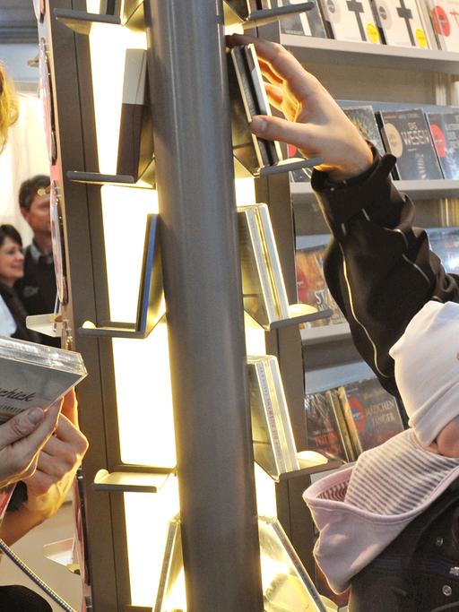 Ein Mann mit einem Kleinkind in einer Tragetasche und eine Frau hören auf der Leipziger Buchmesse mit Kopfhörern Bücher im Bereich Hörbücher.