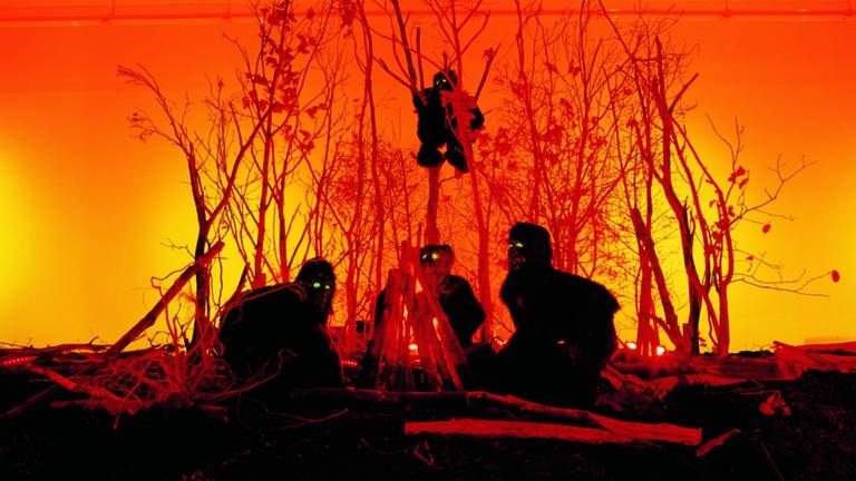 In der Installation "Apes" von Olaf Breuning aus dem Jahr 2001 sitzen menschengroße Affen zwischen hohen Büschen, im Hintergrund ein gelb-roter Himmel wie bei einem Sonnenuntergang.
