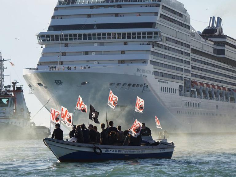 Als Silhouetten heben sich Demonstranten mit Plakaten in einem kleinen Boot vor einem riesigen Kreuzfahrtschiff ab.