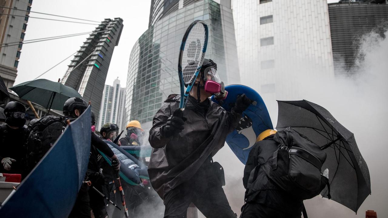 Ein Demonstrat stellt sich mit Helm, Schutzmaske und einem Tennischschläger in der Hand der Polizeit und dem aufkommden Rauch entgegen.