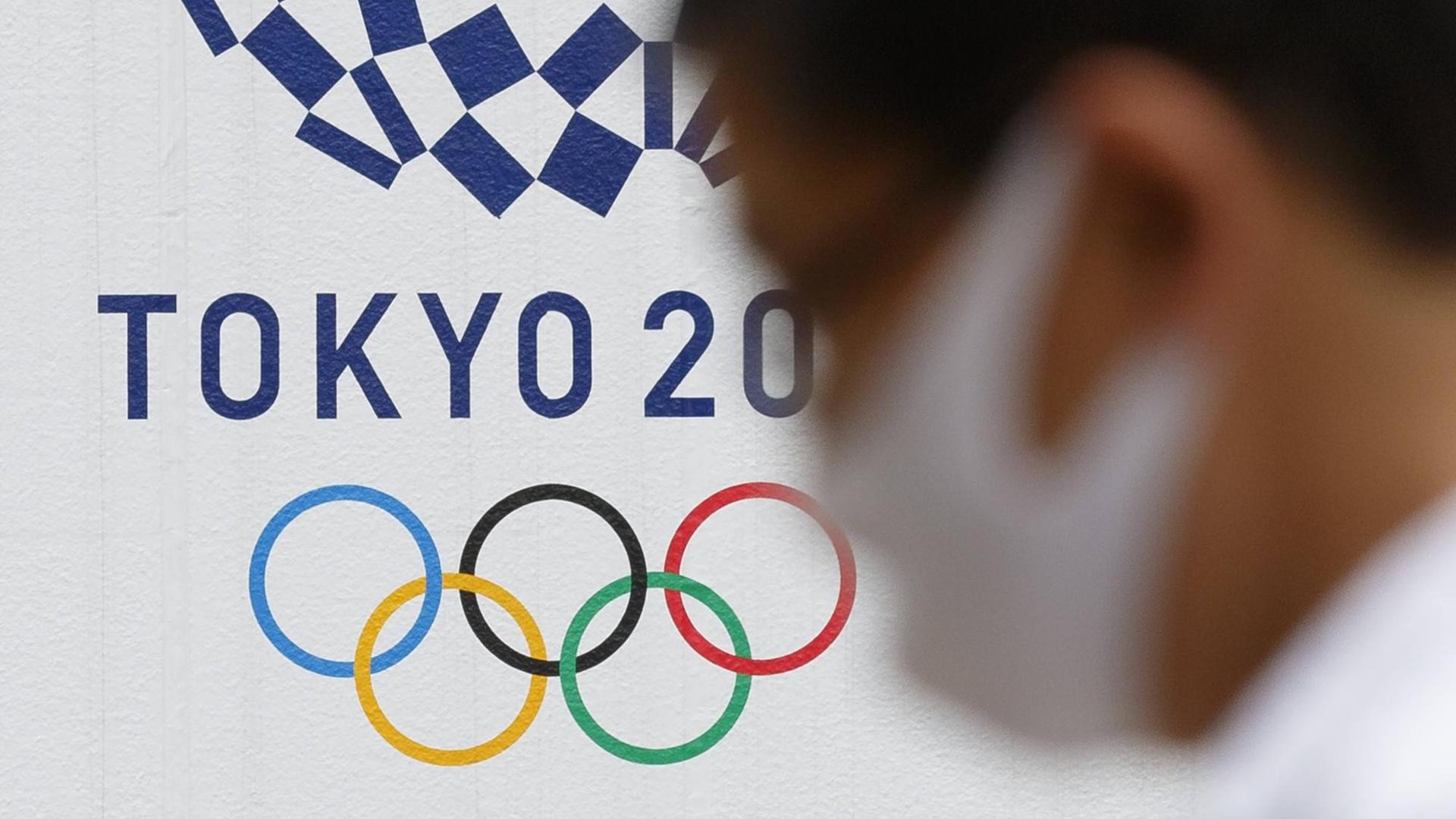 Ein Mann mit Gesichts-Maske geht an einem Logo für die Olympischen Spiele in Tokio vorbei.