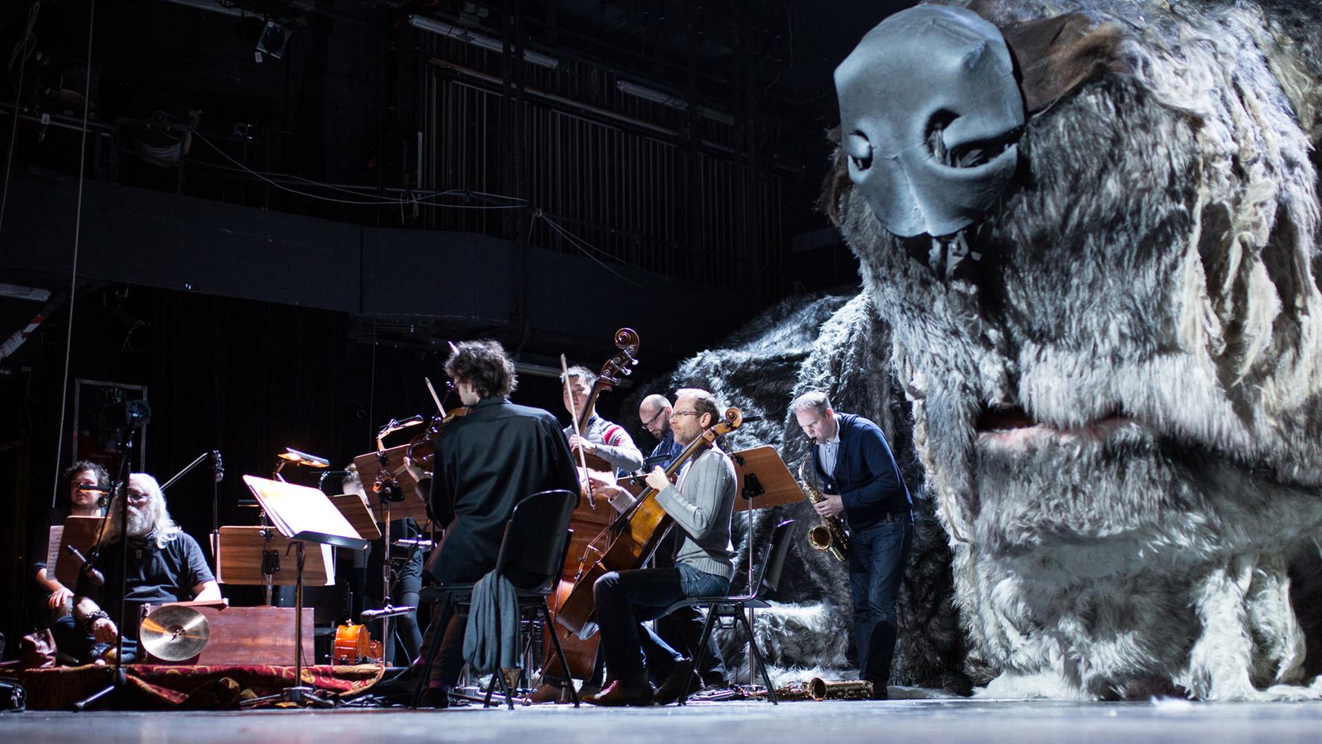 Eine überdimensionale Figur eines Hundes liegt in Bochum im Schauspielhaus während einer Probe auf der Bühne, während Musiker daneben sitzen.