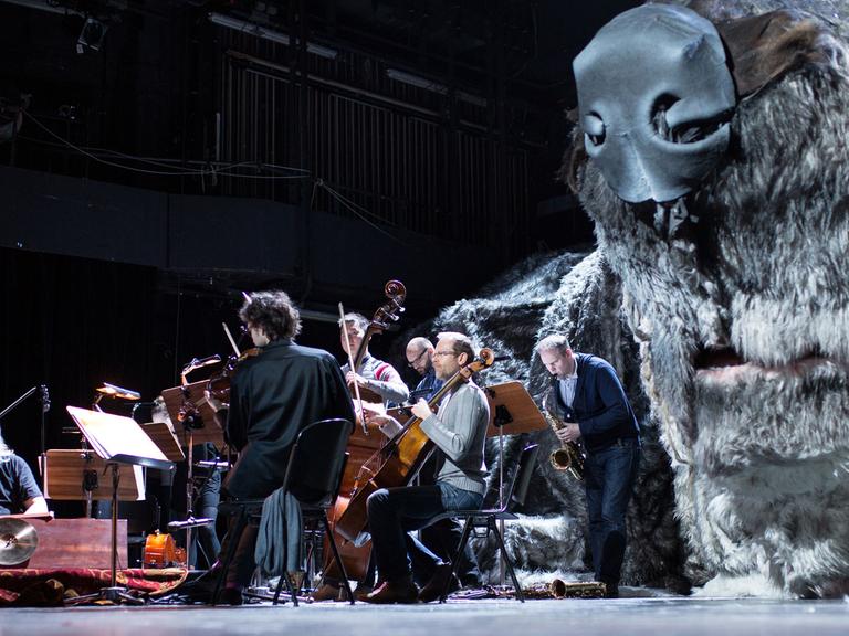 Eine überdimensionale Figur eines Hundes liegt in Bochum im Schauspielhaus während einer Probe auf der Bühne, während Musiker daneben sitzen.