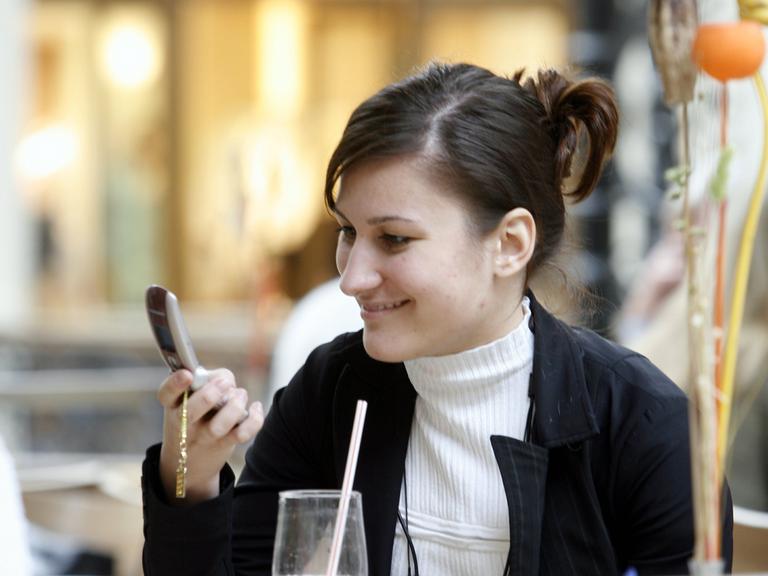 Eine junge Frau schaut auf ihr Mobiltelefon, während sie in einem Cafe sitzt. 