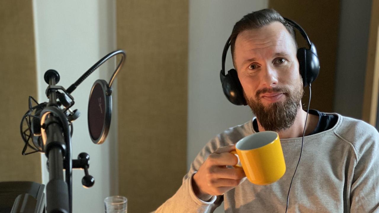 Robert Marc Lehmann am Lesepult vor dem Studio-Mikrofon, mit Kopfhörern auf den Ohren und einer gelben Kaffeetasse in der Hand
