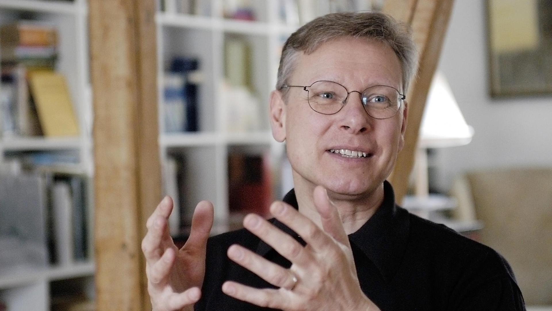 Ein mittelalter Mann mit Brille gestikuliert beim Reden mit den Händen. Im Hintergrund ist ein Bücherschrank zu sehen.