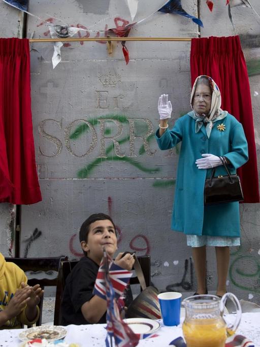 Kinder und eine als Königin Elizabeth verkleidete Person bei einem Event des britischen Künstlers Banksy.