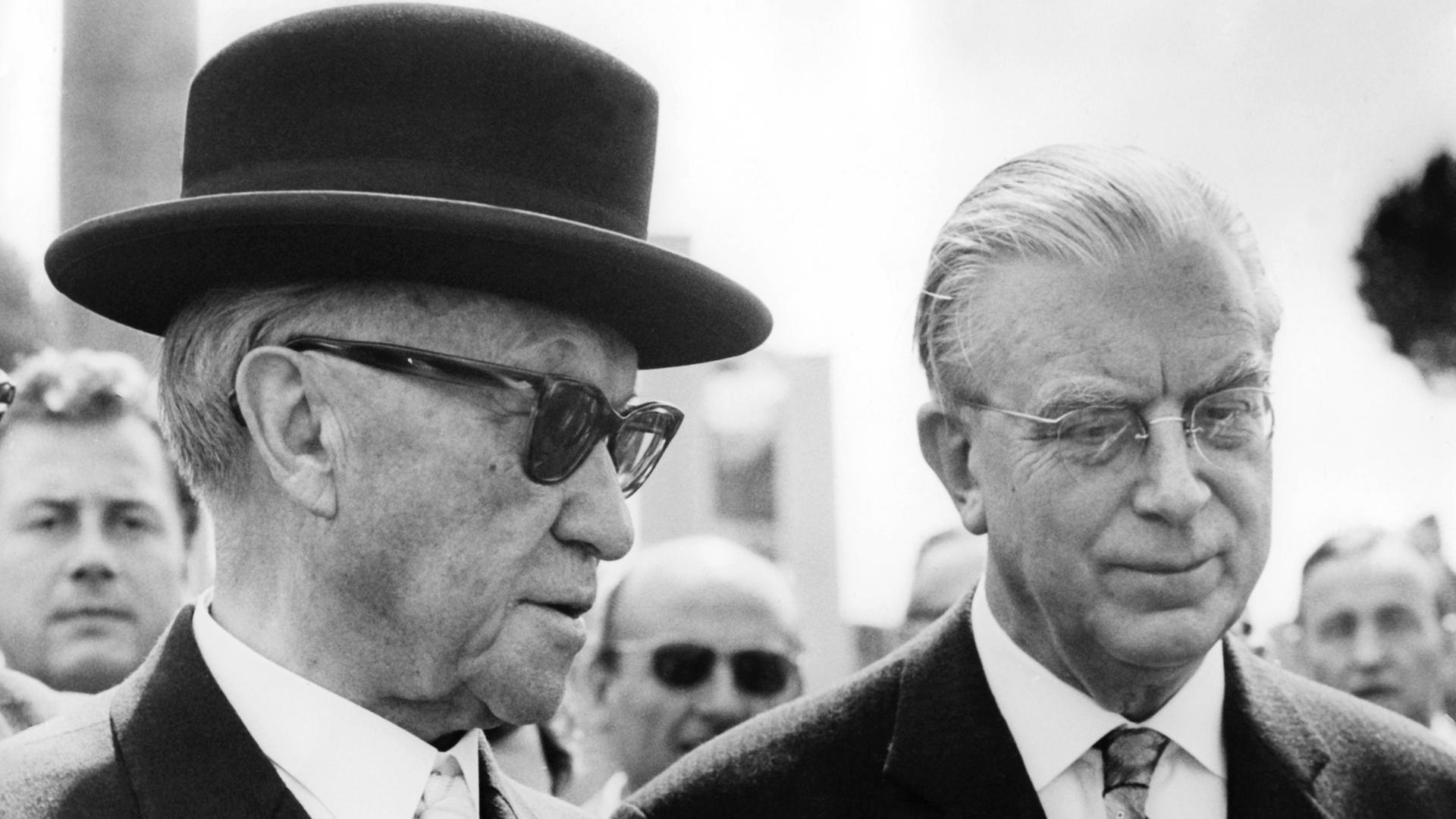 Kanzler Konrad Adenauer trägt eine Sonnenbrille und einen schwarzen Hut, rechts neben ihm steht Kanzleramtschef Hans Globke