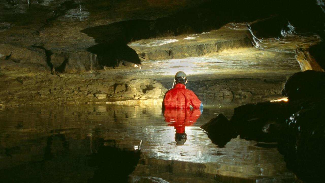 Ein Höhlenforscher in einem roten Anzug durchquert einen unterirdischen See in der Falkensteiner Höhle.