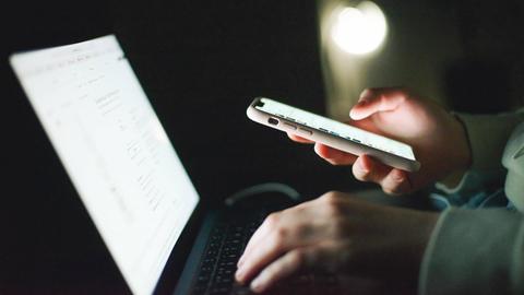 Ein Frau schaut auf einen Laptop und hält zugleich ein eingeschaltetes Smartphone in der Hand.