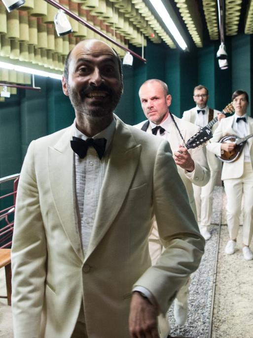 Vier Männer in weißen Anzügen, davon drei mit Saiteninstrumenten, tänzeln in einer Reihe durch einen Konzertsaal