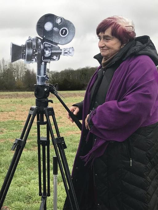 Agnes Varda steht auf einem Kamerawagen der auf Schienen in einer Landschaft steht.