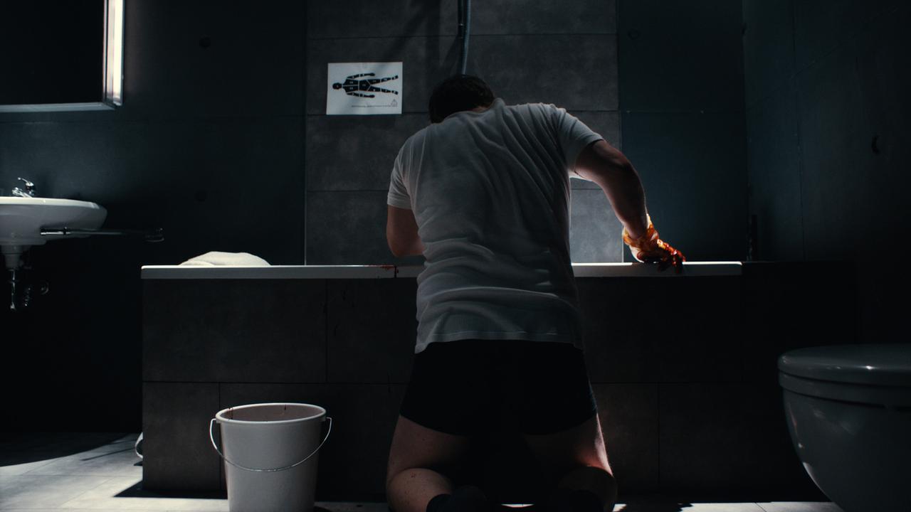 Adam Ild Rohweder kniet in einer Szene von "A Young Man with High Potential" in Unterwäsche mit blutigen Handschuhen vor einer Badewanne. Im Raum ist es dunkel, er wird von hinten gezeigt. Über der Badewanne klebt ein weißer Zettel, auf der in schwarz ein Körper aufgedruckt ist, der in verschiedene Teile unterteilt ist.