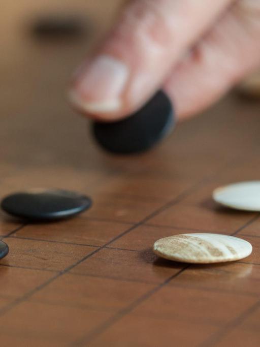 Das Bild zeigt zwei Finger in Nahaufnahme, die einen schwarzen Go-Stein auf dem Spielfeld versetzen.