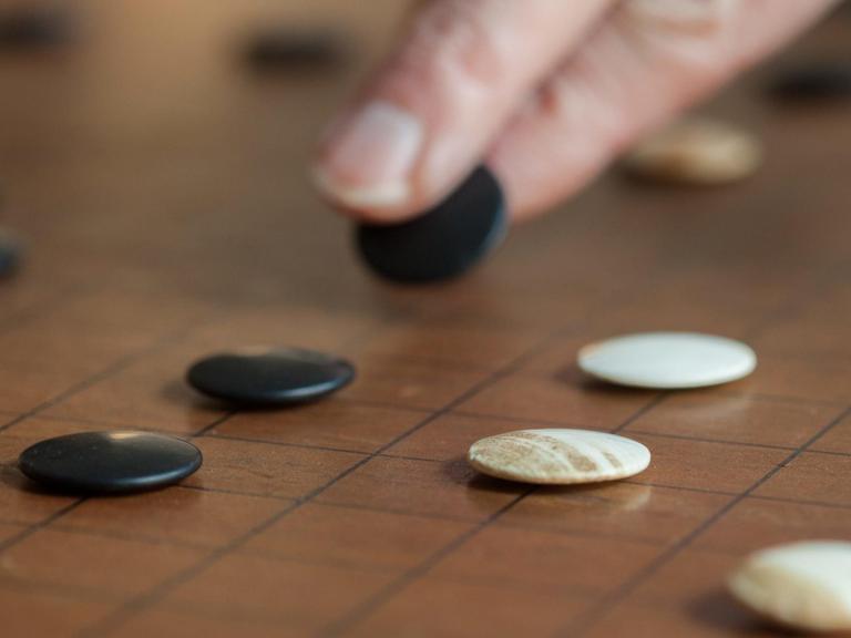 Das Bild zeigt zwei Finger in Nahaufnahme, die einen schwarzen Go-Stein auf dem Spielfeld versetzen.