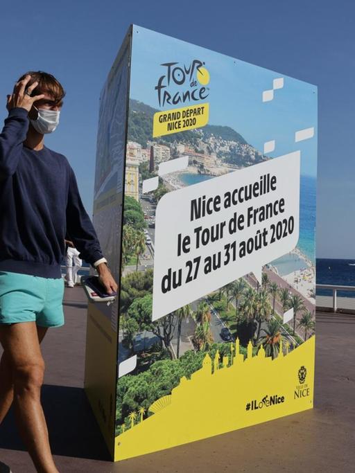 Zwei Passanten mit Mundschutz gehen auf der Promenade des Anglais in Nizza an einem Plakat vorbei, das die Tour de France 2020 ankündigt