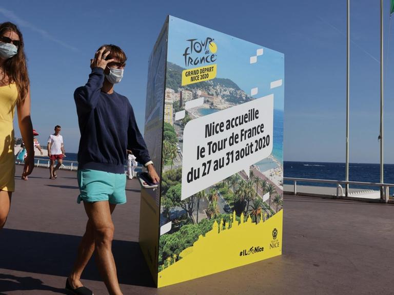 Zwei Passanten mit Mundschutz gehen auf der Promenade des Anglais in Nizza an einem Plakat vorbei, das die Tour de France 2020 ankündigt