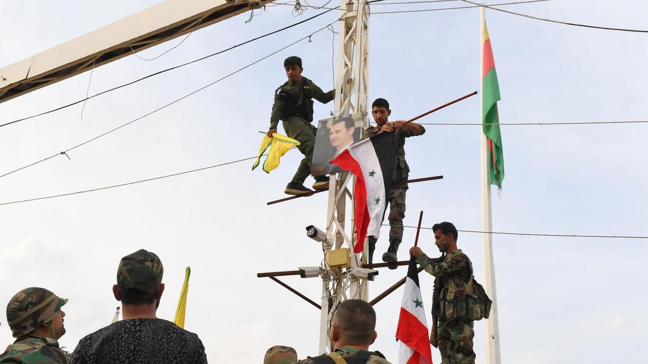 Syrische Regierungssoldaten klettern an der türkischen Grenze in der Stadt Kobane auf einen Strommast mit der Nationalflagge der syrischen Regierung und der gelben Fahne der Kurdischen Volksschutzeinheiten (YPG), zusammen mit einem Porträt des syrischen Präsidenten Bashar al-Assad. Im Hintergrund weht die gelb-rot-grünen kurdischen Rojava-Flagge auf einem Mast.