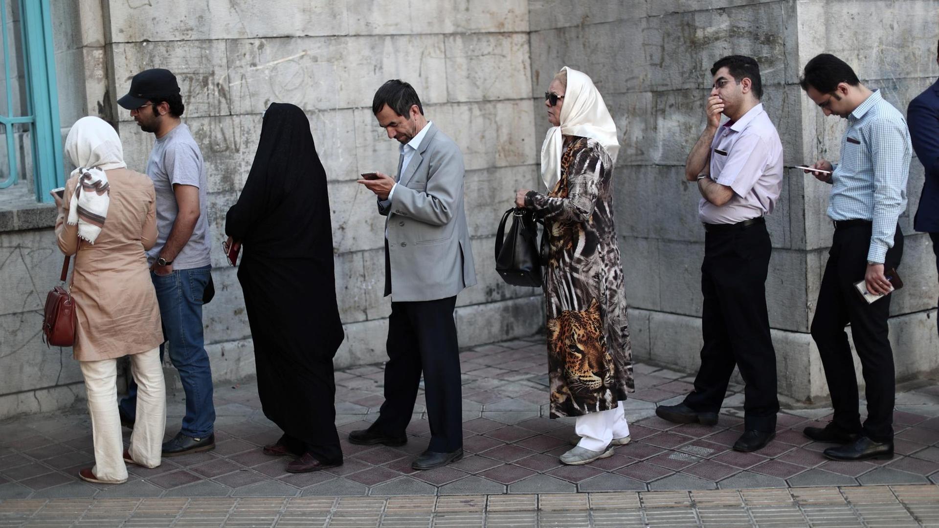 Mehrere Frauen und Männer stehen vor einem Gebäude in einer lockeren Warteschlange. Einige unterhalten sich, andere blicken auf ihre Smartphones.