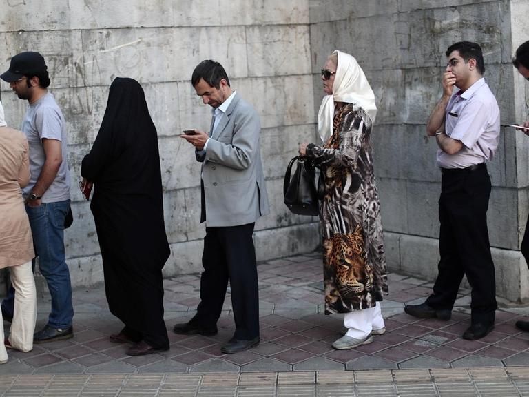 Mehrere Frauen und Männer stehen vor einem Gebäude in einer lockeren Warteschlange. Einige unterhalten sich, andere blicken auf ihre Smartphones.