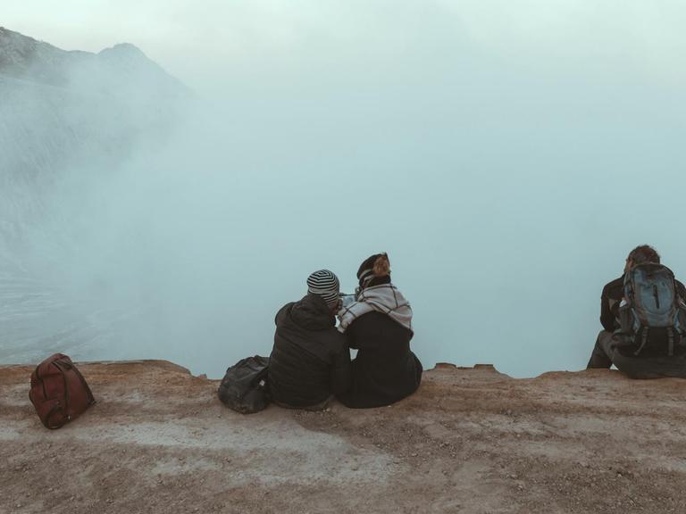 Drei junge Menschen sitzen mit Rucksäcken auf einem Berg und schauen in die Wolken.