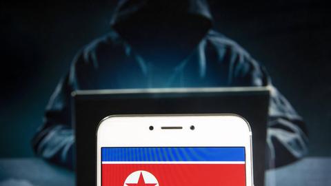 Nordkoreanische Flagge auf Handy vor dunkler Hacker-Gestalt im Kapuzenpulli