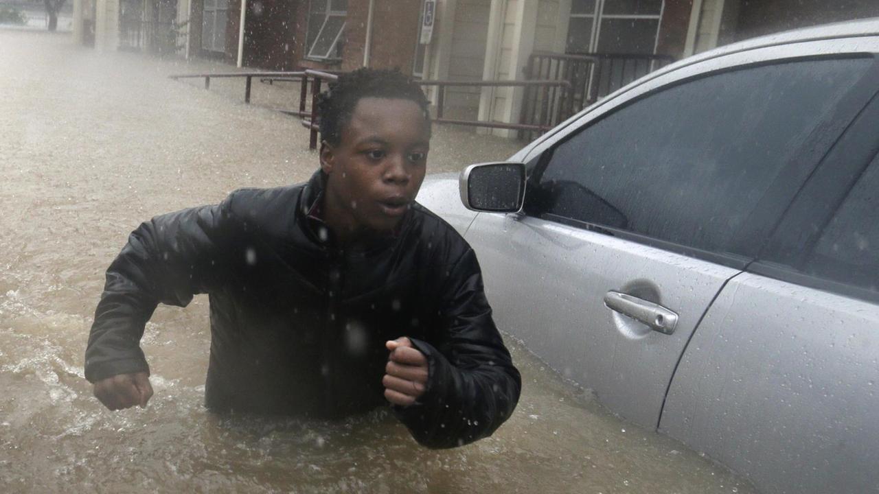 Der Junge läuft in einer Straße neben einem Auto durch das Wasser, das ihm bis über die Gürtellinie reicht. Es regnet in Strömen.