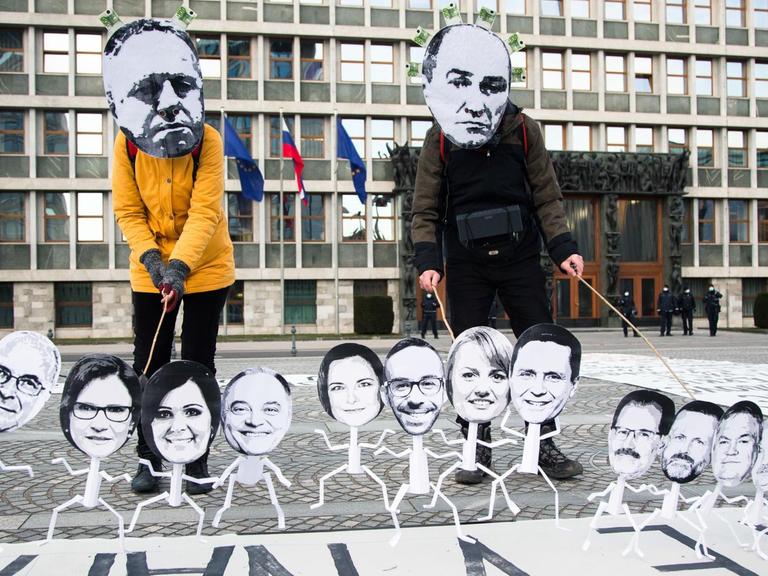 Zwei Menschen tragen Masken mit Ministerpräsident Janez Janša und dem Wirtschaftsminister Zdravko Pocivalsekâ. Sie wiederum halten kleine Figuren an Stöcken, es sind die anderen Mitglieder des slowenischen Parlaments.