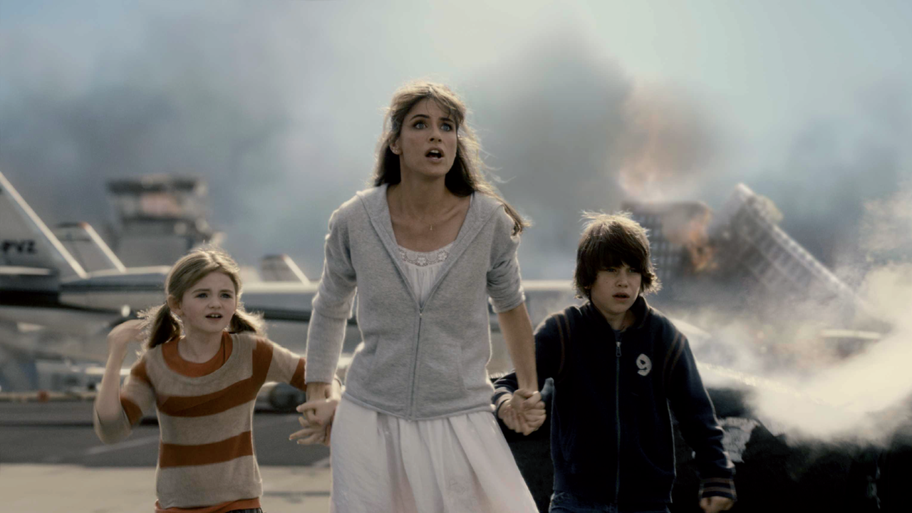 Eine Frau hält an ihren Händen zwei Kinder. Sie schauen sehr erschrocken. Hinter ihnen steht ein Flugzeug und ein brennendes, eingestürztes Gebäude. Es handelt sich um eine Szene aus dem Katastropenhfilm "2012" von Roland Emmerich.